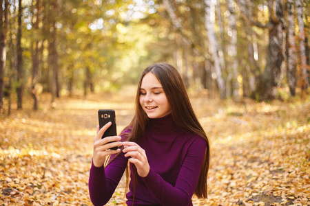 可爱的十几岁女孩带智能手机。秋天森林里欢快的女孩的肖像。