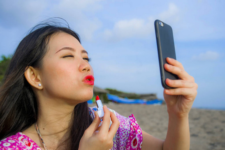 年轻快乐美丽的亚洲女性在暑假旅行期间，在热带天堂海滩用手机当镜子，在嘴唇上涂红色口红，修饰自己的妆容