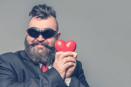 成年留胡子的男人穿着西装，戴着太阳镜，在灰色背景上抱着一颗红色的心。孤立