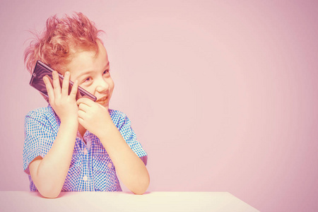 可爱的小男孩在一个粉红色的背景下坐在桌子旁打电话。划