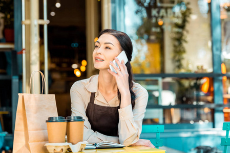 漂亮的女服务员，穿着围裙，坐在桌子上，在咖啡馆附近的街道上用智能手机说话