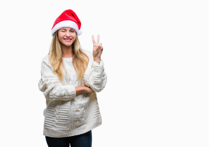 年轻漂亮的金发女人戴着圣诞帽，在孤立的背景上微笑，快乐的脸对着镜头眨着眼睛，做着胜利的标志。 二号。