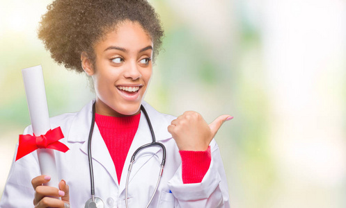 年轻的AFRO美国医生女士拿着学位，在孤立的背景上指着，用拇指向侧面显示，脸上带着快乐的微笑