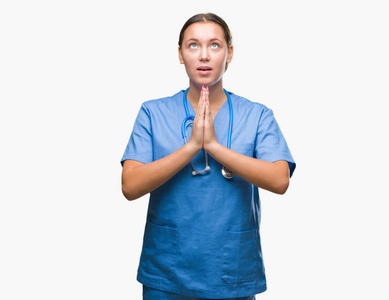 年轻的白种人医生女士穿着医疗制服，在孤立的背景下乞讨和祈祷，双手一起祈祷，脸上充满希望的表情，非常情绪化和担忧。请求原谅。宗教观