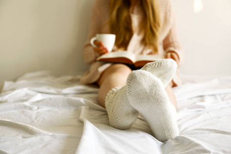 懒洋洋的下午概念..在晨光中，坐在床上的女人靠在白色的墙上，在窗边读着旧书，喝着一杯卡布奇诺咖啡。背景，复制空间