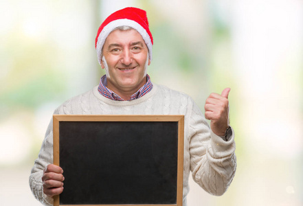 英俊的老人戴着圣诞帽，拿着黑板在孤立的背景上指指点点，用拇指指向一边，脸上洋溢着幸福的笑容