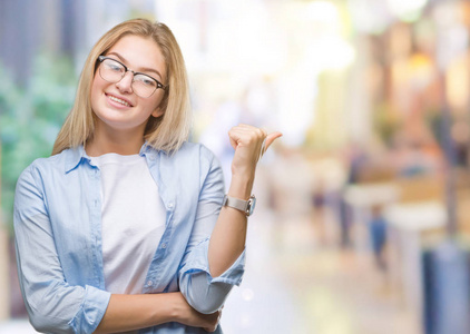 年轻的白种人商业女性戴着眼镜，戴着孤立的背景，微笑着，快乐的脸看着，拇指向上指向一边。