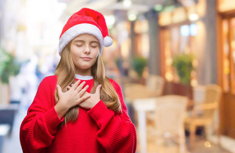 年轻漂亮的女孩戴着圣诞帽在孤立的背景上微笑，双手放在胸前，闭着眼睛，脸上有感激的手势。健康观念。