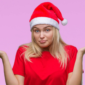 年轻的白种人女人戴着圣诞帽，在孤立的背景上毫无头绪，表情混乱，举起手臂和手。怀疑的概念。