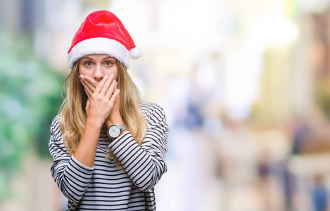 年轻漂亮的金发女人戴着圣诞帽，在孤立的背景下震惊地用手捂住嘴，因为错误。秘密概念。