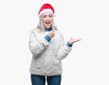 年轻的金发女人戴着圣诞帽在孤立的背景上惊讶和微笑的相机,同时呈现