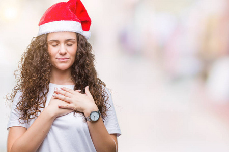年轻的黑发女孩戴着圣诞帽，在孤立的背景上微笑，双手放在胸部，闭着眼睛，脸上有感激的手势。 健康概念。