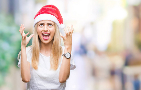 年轻漂亮的金发女人圣诞帽在孤立的背景上疯狂和疯狂的喊叫和喊叫，咄咄逼人的表情和手臂举起。 挫折概念。