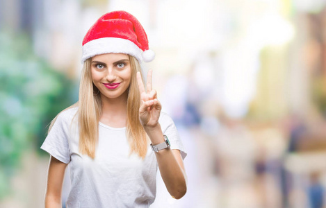 年轻漂亮的金发女人圣诞帽在孤立的背景上微笑，快乐的脸对镜头眨眼，做胜利标志。 二号。