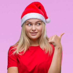 年轻的高加索女人戴着圣诞帽，在孤立的背景上微笑，快乐的脸看着，用拇指指向一边。