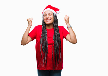 年轻的辫子非洲裔美国女孩戴着圣诞帽，在孤立的背景上庆祝，惊讶和惊讶的成功，举起手臂和睁开眼睛。 赢家概念。