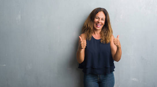中年西班牙裔妇女站在灰色的格朗格墙上，成功的标志，用手竖起大拇指，微笑和快乐地做积极的手势。 用欢快的表情看着相机，胜利者的手势