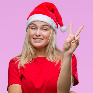年轻的高加索女人戴着圣诞帽，在孤立的背景上微笑，快乐的脸对着镜头眨着眼睛，做着胜利的标志。 二号。