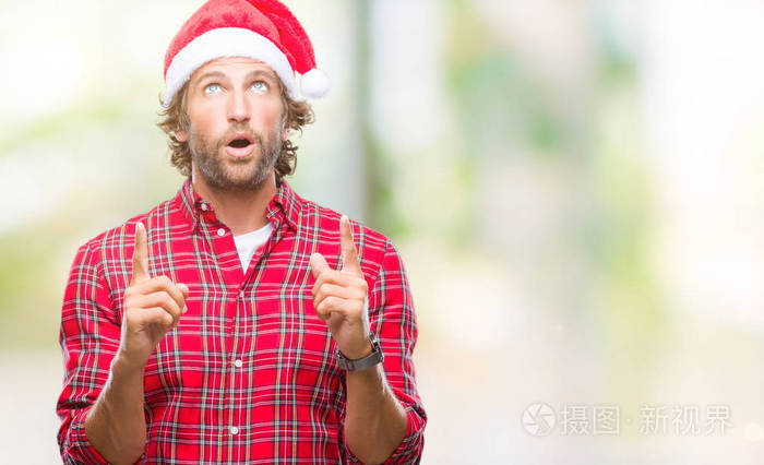 英俊的西班牙男子模特穿着圣诞老人的圣诞老人在孤立的背景上惊讶和惊讶地抬头，用手指和举起的手臂指着。