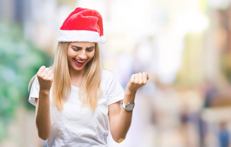 年轻漂亮的金发女人圣诞帽在孤立的背景下，非常高兴和兴奋地做胜利者的手势，举起手臂，微笑和尖叫，争取成功。 庆祝概念。