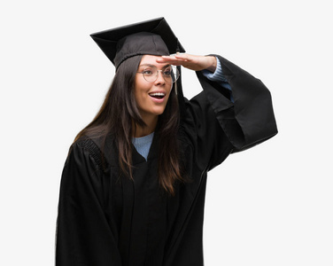 年轻的西班牙裔女人戴着毕业帽和制服，非常高兴和微笑，用手捂住头看着远方。 搜索概念。
