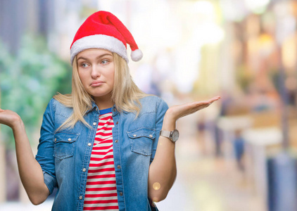 年轻的高加索女人戴着圣诞帽，在孤立的背景上毫无头绪，表情混乱，举起手臂和手。 怀疑概念。