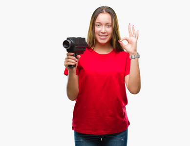 年轻美丽的白种人女人用老式摄像机在孤立的背景上拍摄，用手指做OK标志，优秀的符号