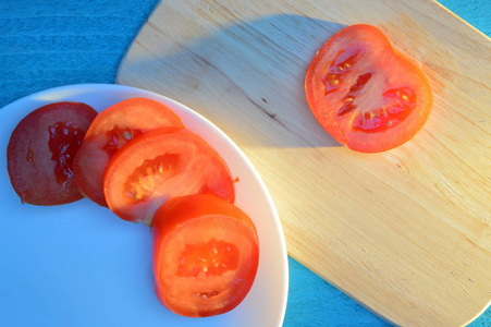 番茄片放在木切割板和白色陶瓷板上，上面有一张蓝色的绿松石桌子，阳光照射下，有长长的冷影