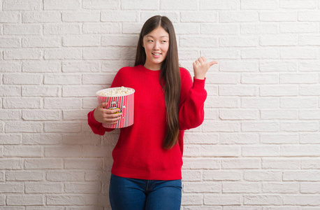 年轻的中国妇女在砖墙上吃着爆米花，用拇指指着一边，脸上洋溢着幸福的笑容