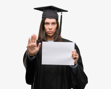 年轻的西班牙裔妇女，穿着毕业制服，张开手拿着文凭纸，做着停止标志，严肃而自信地表达着防御手势
