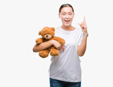 年轻的白种人女人抱着泰迪熊，在孤立的背景下惊讶地发现了一个想法或问题，手指着幸福的脸第一