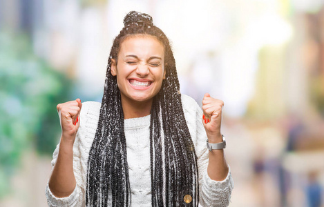 年轻的辫子非洲裔美国女孩穿着毛衣，在孤立的背景下兴奋地成功，举起双臂庆祝胜利，微笑。 赢家概念。