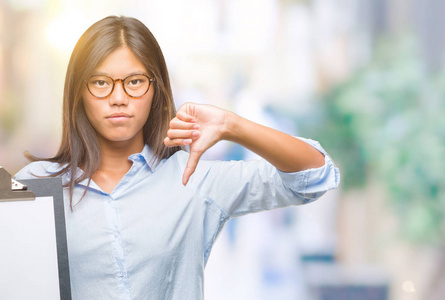 年轻的亚洲商界女性，在孤立的背景下，拿着剪贴板，愤怒的脸，负面的标志，表示不喜欢，竖起大拇指拒绝概念