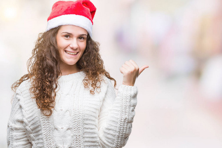 年轻的黑发女孩戴着圣诞帽，在孤立的背景上微笑，快乐的脸看着，用拇指指向一边。