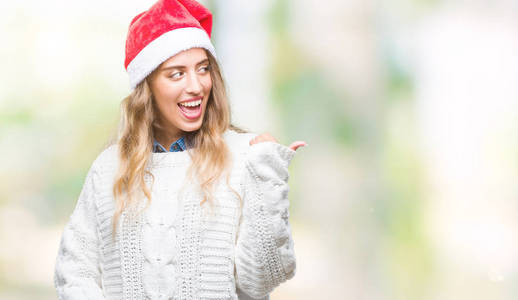 美丽的年轻金发女人戴着圣诞帽，在孤立的背景上微笑，快乐的脸看着，用拇指指向一边。