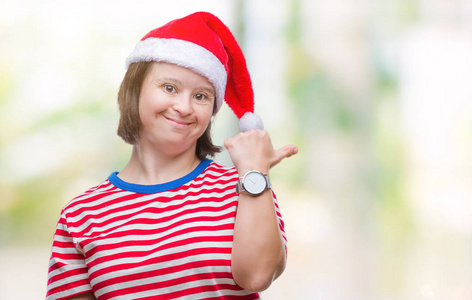 年轻的成年妇女患有唐氏综合症，戴着圣诞帽，在孤立的背景上微笑，快乐的脸看着，拇指向上指向一边。