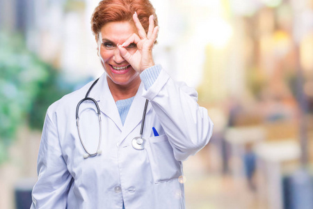 高级白种人医生女士穿着医疗制服，在孤立的背景下做着OK的手势，用手微笑的眼睛透过手指看着幸福的脸。
