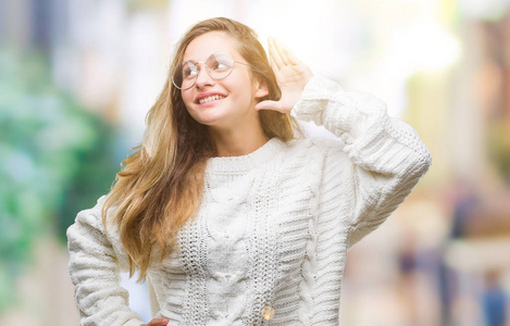 年轻漂亮的金发女人穿着冬天的毛衣和太阳镜，在孤立的背景上微笑，用手捂住耳朵听谣言或流言蜚语。 耳聋概念。