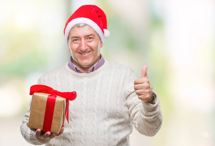 英俊的老人戴着圣诞帽，抱着礼物在孤立的背景上，开心地大笑着，做着，好的，用手指签个字，大拇指就好了