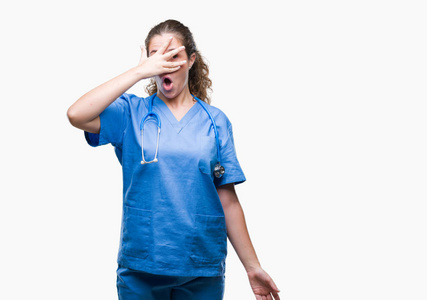 年轻的黑发医生女孩穿着护士或外科医生的制服，在孤立的背景上偷看震惊，覆盖着脸和眼睛，用手透过手指看着尴尬的表情。