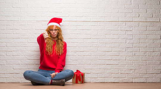 年轻的红头发妇女坐在砖墙上戴着圣诞帽，看上去不高兴和愤怒，表现出拒绝和消极的拇指向下的手势。 不好的表情。