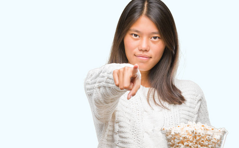 年轻的亚洲女人在孤立的背景上吃爆米花，手指指向相机，并指向你的手，从前面签署积极和自信的手势。