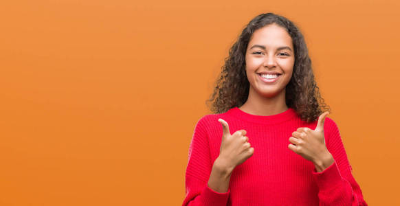 年轻的西班牙裔妇女穿着红色毛衣，成功标志，用手竖起大拇指，微笑和快乐地做积极的手势。 用欢快的表情看着相机，胜利者的手势。
