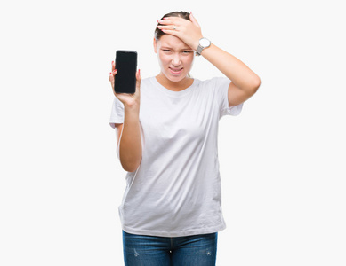 年轻漂亮的高加索女人在孤立的背景上显示智能手机屏幕，用手压着头，羞愧和惊讶的脸，愤怒和沮丧。 因为错误而恐惧和不安。