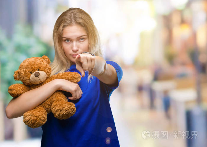 年轻的白种人女人拿着可爱的泰迪熊在孤立的背景上，手指着相机，向你手势正面和自信的手势。