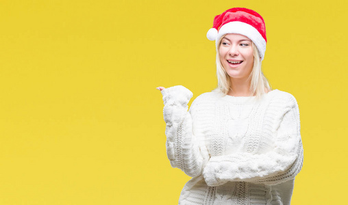 年轻漂亮的金发女人戴着圣诞帽，在孤立的背景上微笑，快乐的脸看着，用拇指指向一边。
