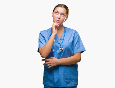 年轻的白种人医生女人穿着医疗制服，在孤立的背景上，手在下巴上思考问题，沉思的表情。 带着体贴的脸微笑。 怀疑概念。
