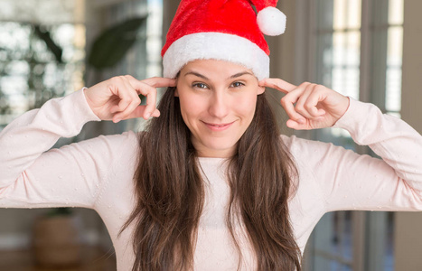 美丽的年轻女子戴着圣诞老人的帽子，在家里用手指遮住耳朵，因为嘈杂的音乐声，她的表情很生气。 聋的概念。