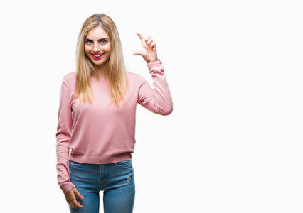 年轻漂亮的金发女人穿着粉红色的冬季毛衣，在孤立的背景上微笑和自信的手势，用手做尺寸标志，同时看和相机。 测量概念。