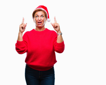 迷人的高级白种人红发妇女戴着圣诞帽，在孤立的背景下，惊讶和惊讶地抬头，用手指和举起的手臂指着。